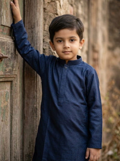 Boy in a navy blue jacquard kurta shalwar set, one hand resting on a rustic wooden door, standing against a textured stone wall background